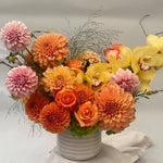 Load image into Gallery viewer, A vibrant floral arrangement featuring oranges, , pinks, and  yellows in a ribbed white ceramic pot, set against a neutral light gray background.
