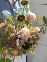 Load image into Gallery viewer, A person in a light blue denim shirt holds a large, rustic floral arrangement featuring sunflowers, pink dahlias, peach roses, and green foliage against a white wall.
