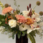 Load image into Gallery viewer, A rustic floral arrangement featuring white roses, peach chrysanthemums, white orchids, and green foliage, presented in a dark amber glass vase against a neutral light gray background.

